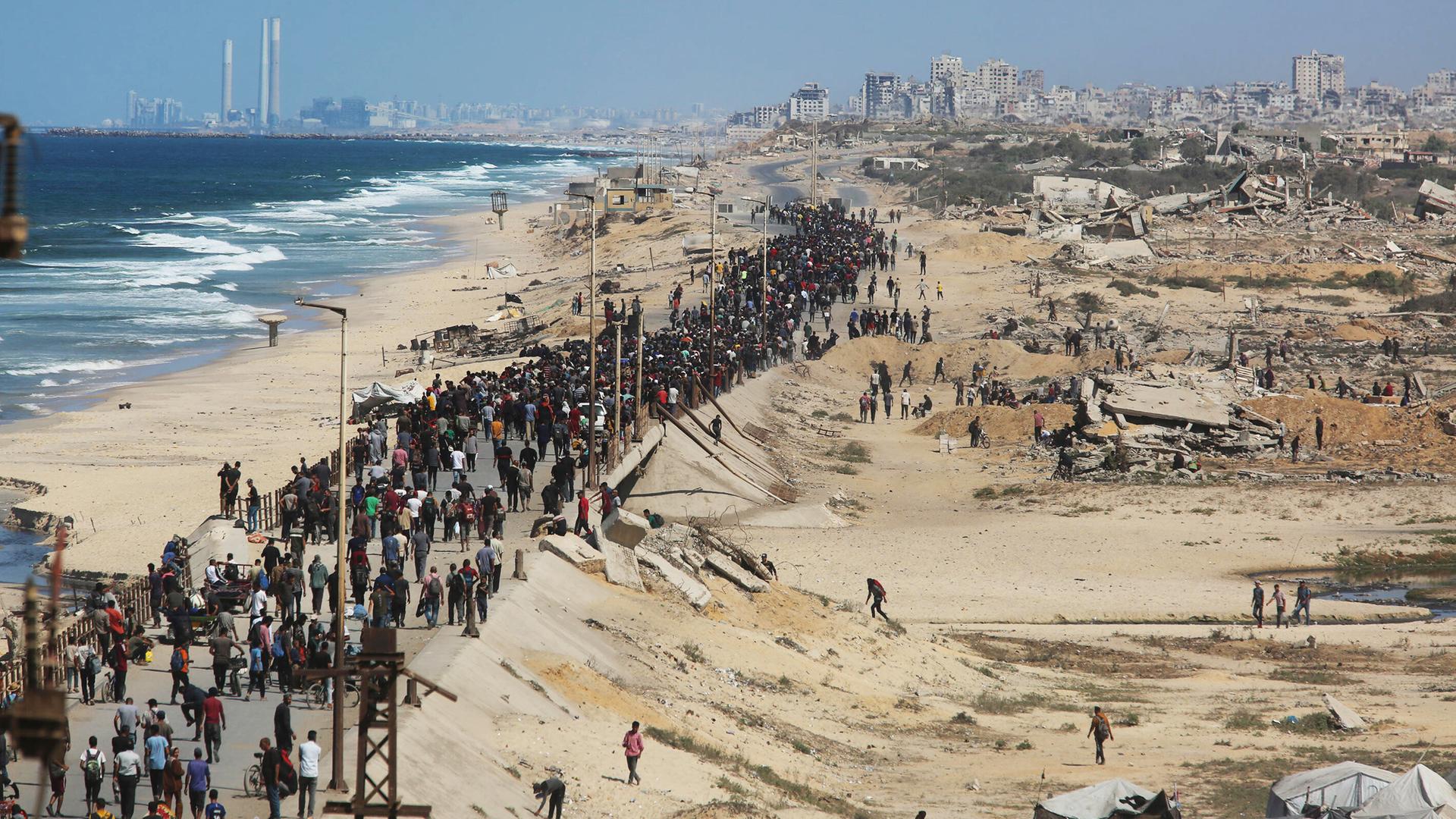 Eine breite Schlange von Menschen läuft einen Standstreifen inmitten des zerstörten Gazastreifens, links das Meer, hinten Ruinen von Gaza-Stadt.