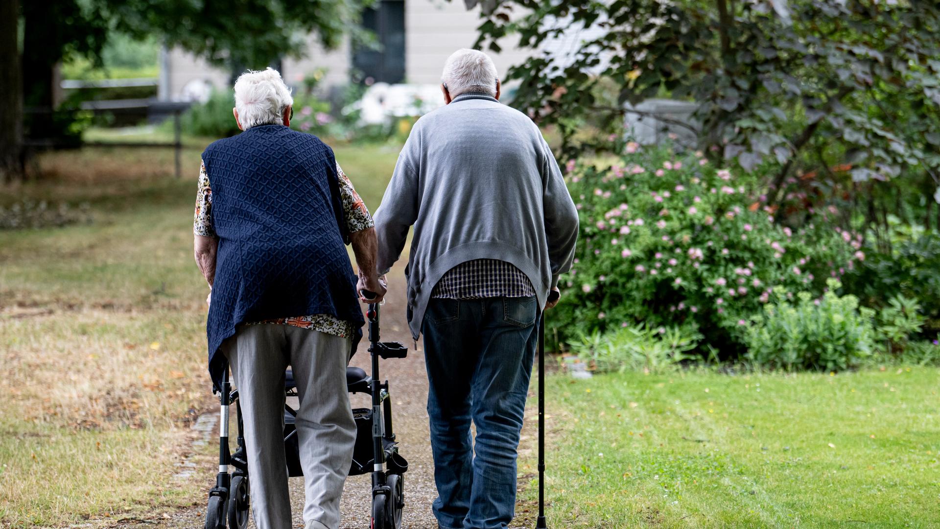 Zwei Senioren gehen mit Gehhilfen durch den Garten eines Seniorenheims. Eine Person nutzt einen Rollator, die andere einen Gehstock. Zwei Senioren gehen mit Gehhilfen durch den Garten eines Seniorenheims. Eine Person nutzt einen Rollator, die andere einen Gehstock.