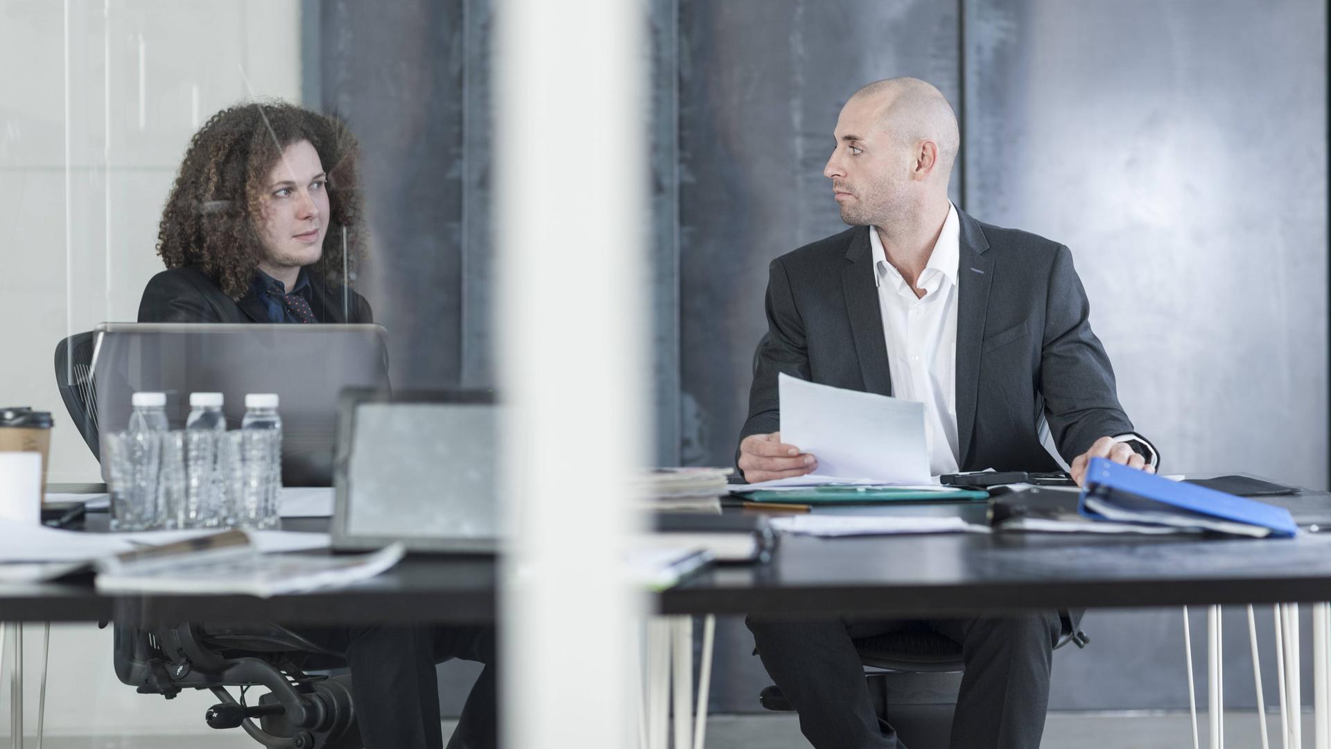 Zwei Personen im Business-Dress - eine jung, die andere etwas älter - sitzen an Schreibtischen und schauen sich skeptisch an.