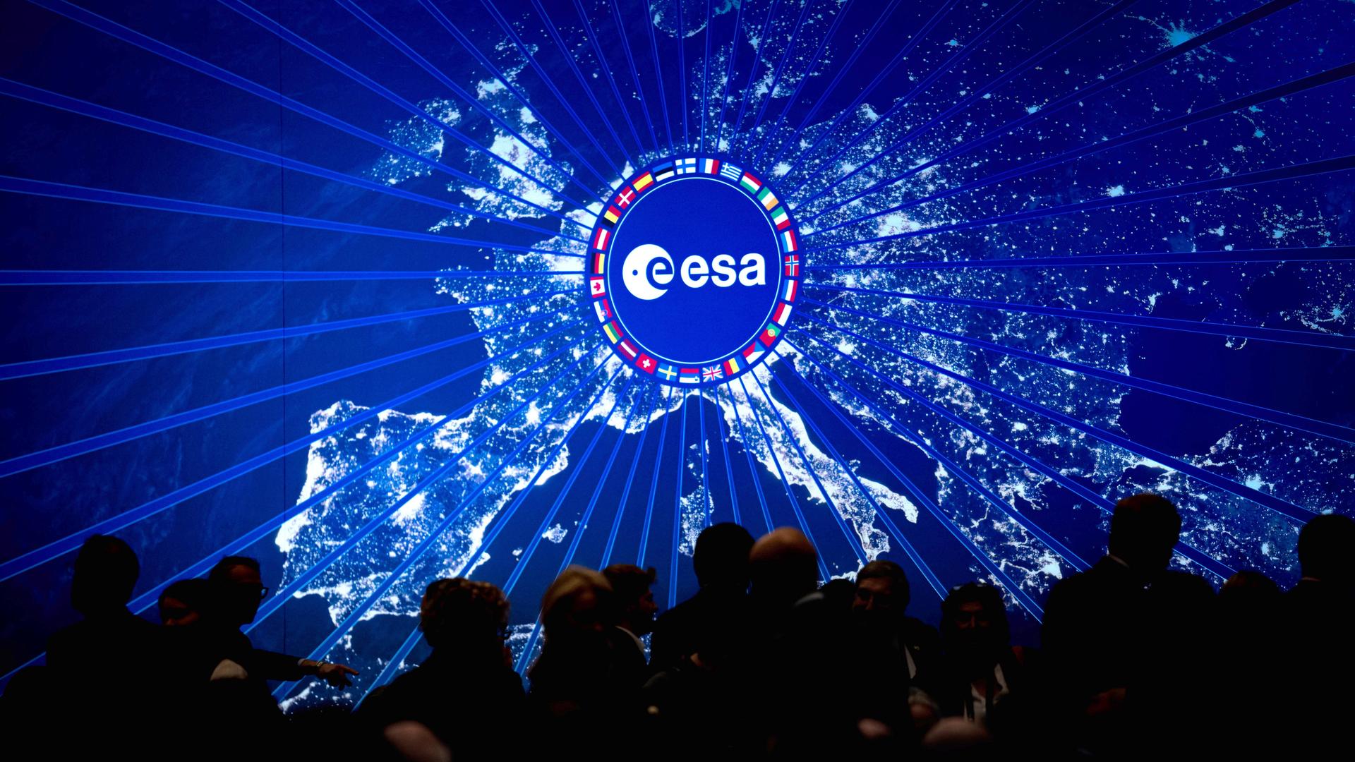Digitale Darstellung Europas bei Nacht mit hell leuchtenden Städten. In der Mitte befindet sich ein rundes Logo der Europäischen Weltraumorganisation (ESA), umgeben von einem Kreis aus Länderflaggen.
