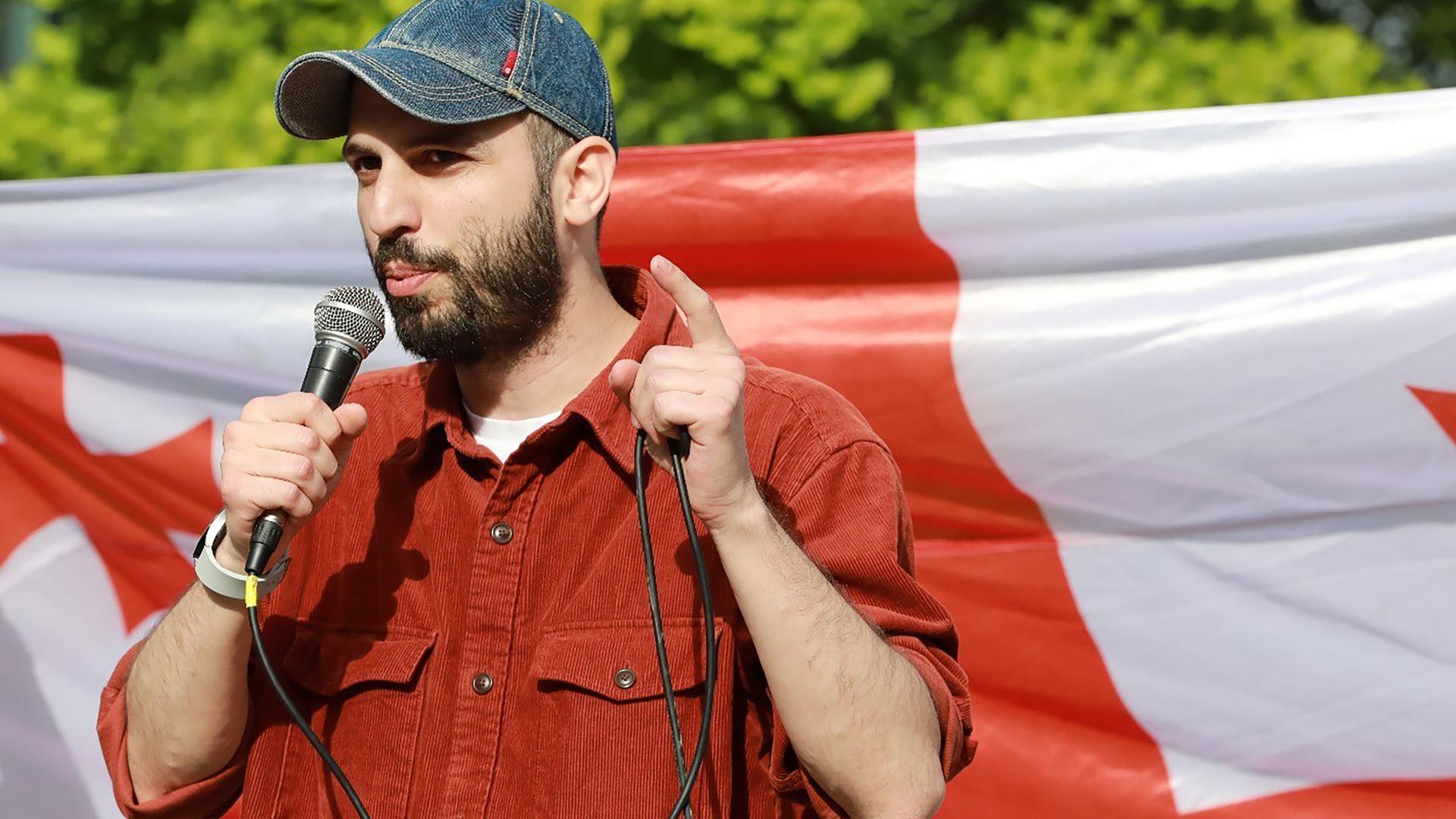 Giorgi Kakabadze steht mit einem Mikrofon in der Hand vor einer georgischen Flagge und spricht Giorgi Kakabadze steht mit einem Mikrofon in der Hand vor einer georgischen Flagge und spricht