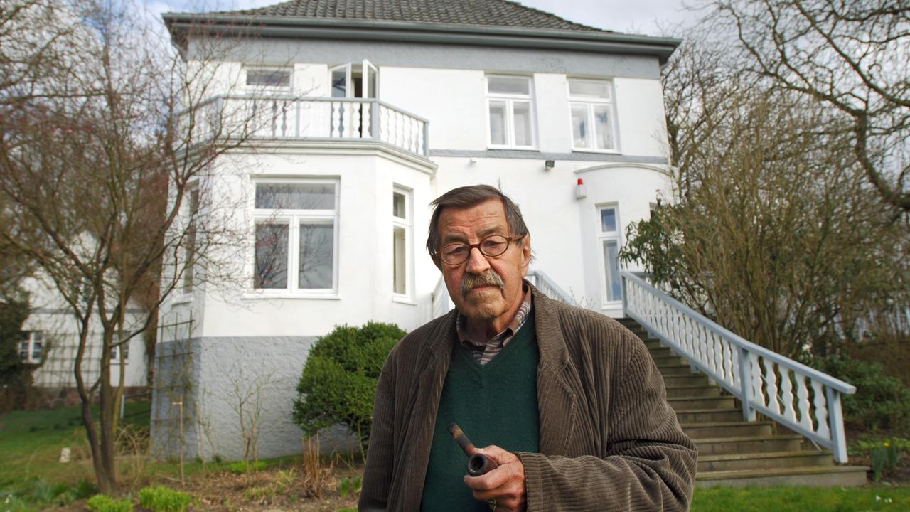 Günter Grass steht im Garten vor seinem Haus in Behlendorf. Grass hält eine Pfeife in der Hand. Das Haus ist zweistöckig. Eine Treppe führt aus dem Garten zum Haus. 