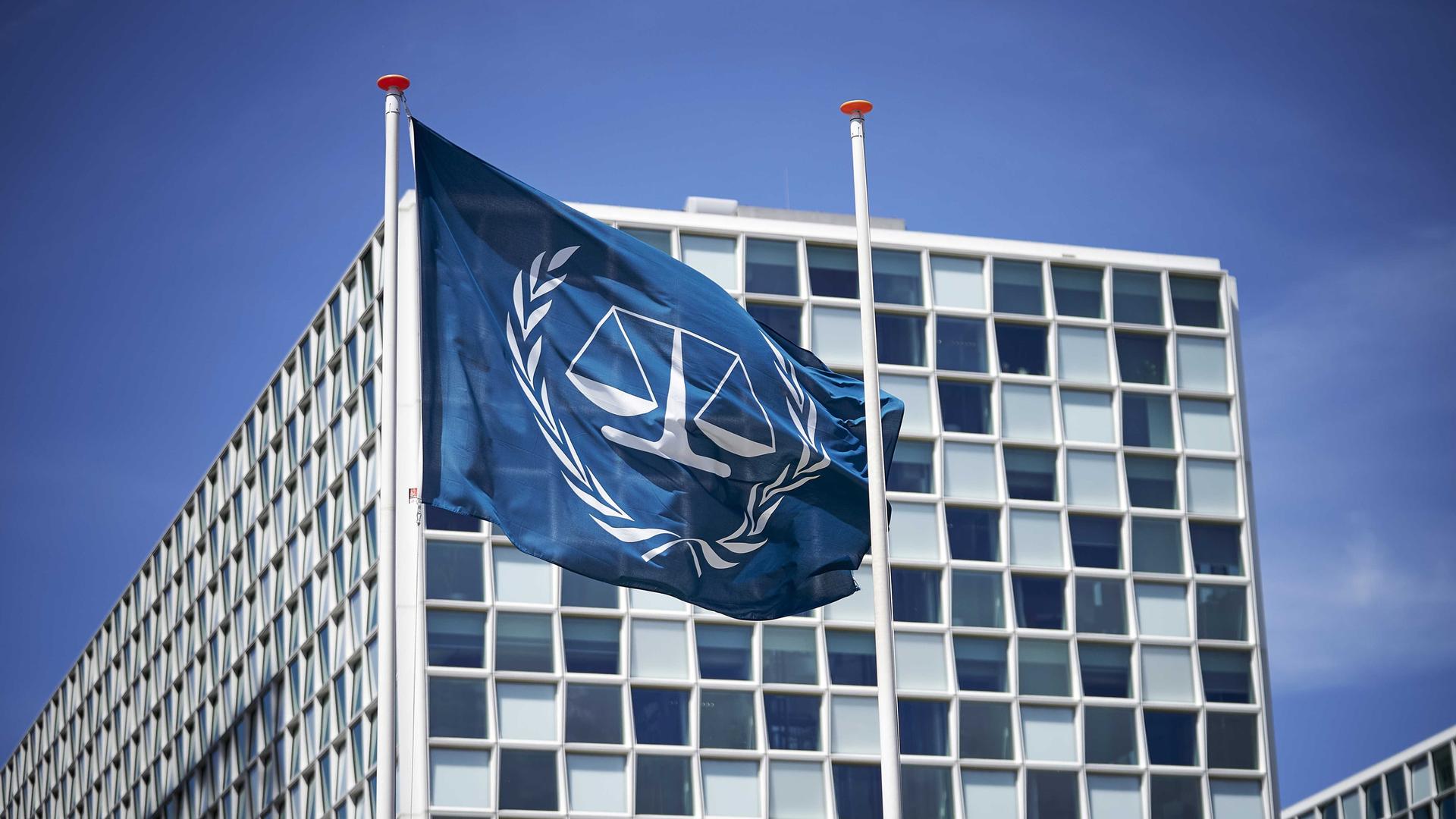 Außenansicht des Internationalen Strafgerichtshofs in Den Haag. Davor eine blaue Flagge, die in weiß das Logo des Gerichts abbildet: eine Waage, die von einem Lorbeerkranz umgeben ist.