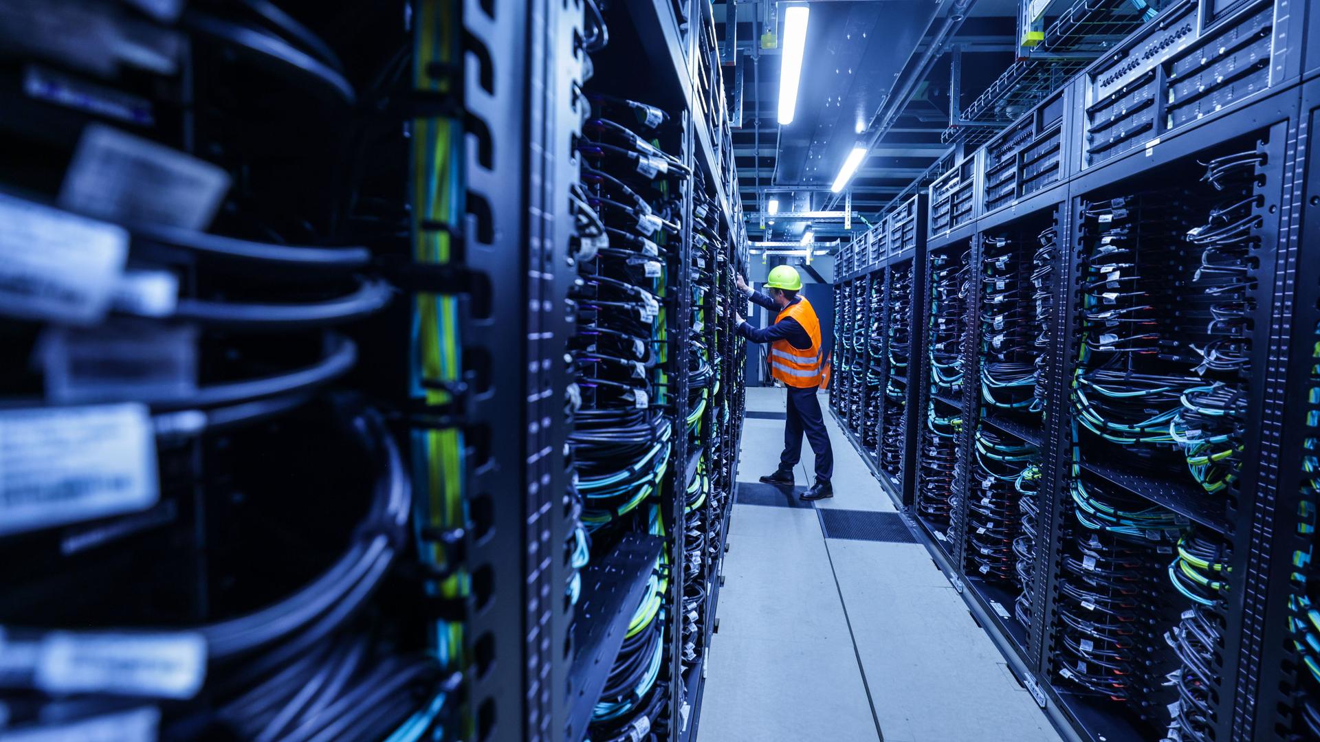 Ein Mitarbeiter mit orange farbener Warnweste iund Sicherheitshelm arbeitet in einem Container des Supercomputers Jupiter im Forschungszentrum Jülich.