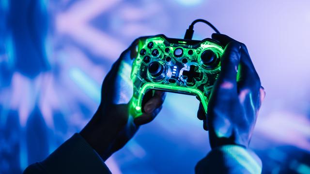 Nahaufnahme zweier Hände die eine Gamingkonsole halten. Die Umgebung ist geprägt von neonfarbenen Lichtern.