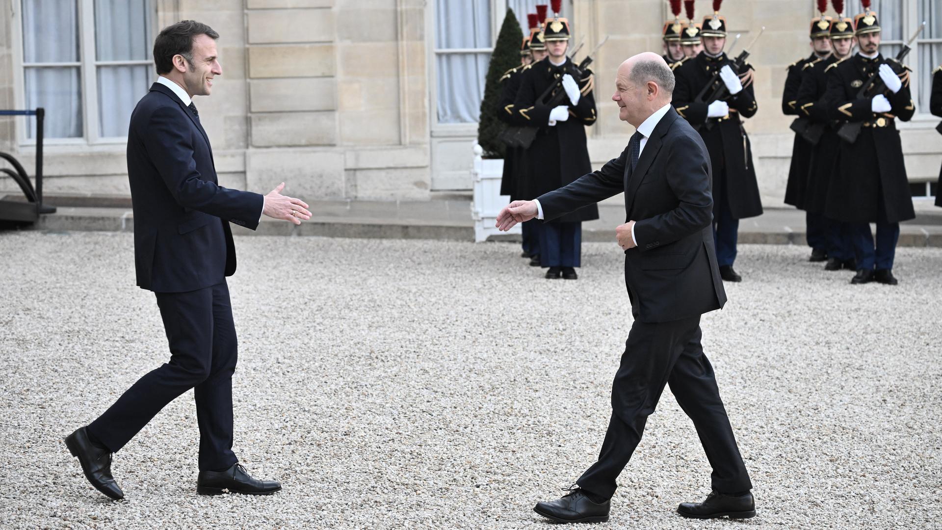 Emmanuel Macron geht mit ausgestreckter Hand auf Olaf Scholz zu. Im Hintergrund stehen französische Soldaten. Emmanuel Macron geht mit ausgestreckter Hand auf Olaf Scholz zu. Im Hintergrund stehen französische Soldaten.