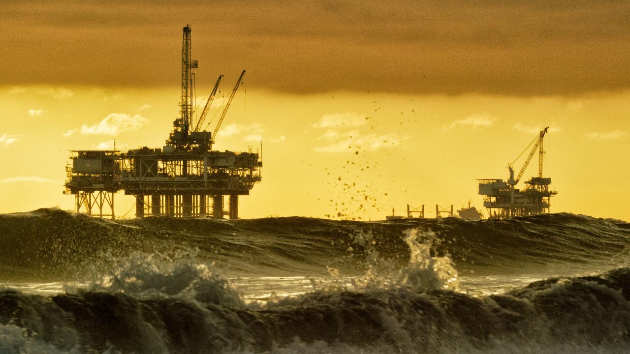 Starke Wellen brechen in der Nähe von Huntington Beach in Südkalifornien, mit mehreren Silhouetten von Offshore-Ölbohrplattformen und einem Öltanker am Horizont in der Ferne bei Sonnenuntergang unter einem dramatischen, stürmischen Himmel.