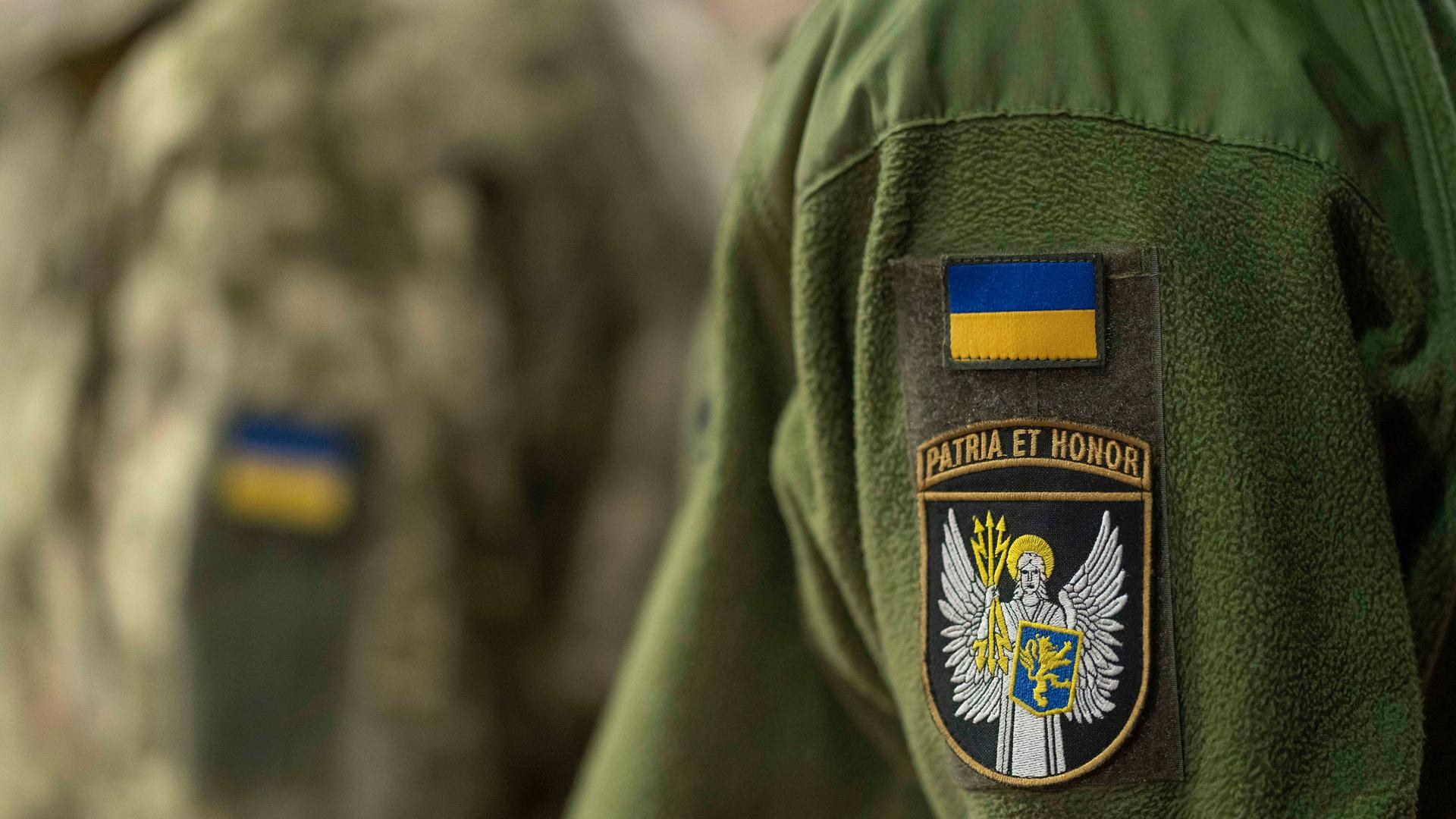 Zwei Personen tragen eine grünfarbene Uniform der ukrainischen Armee. Nur die Jacken sind zu sehen. Auf beiden ist je ein Aufnäher mit der ukrainischen Flagge genäht. Zwei Personen tragen eine grünfarbene Uniform der ukrainischen Armee. Nur die Jacken sind zu sehen. Auf beiden ist je ein Aufnäher mit der ukrainischen Flagge genäht.