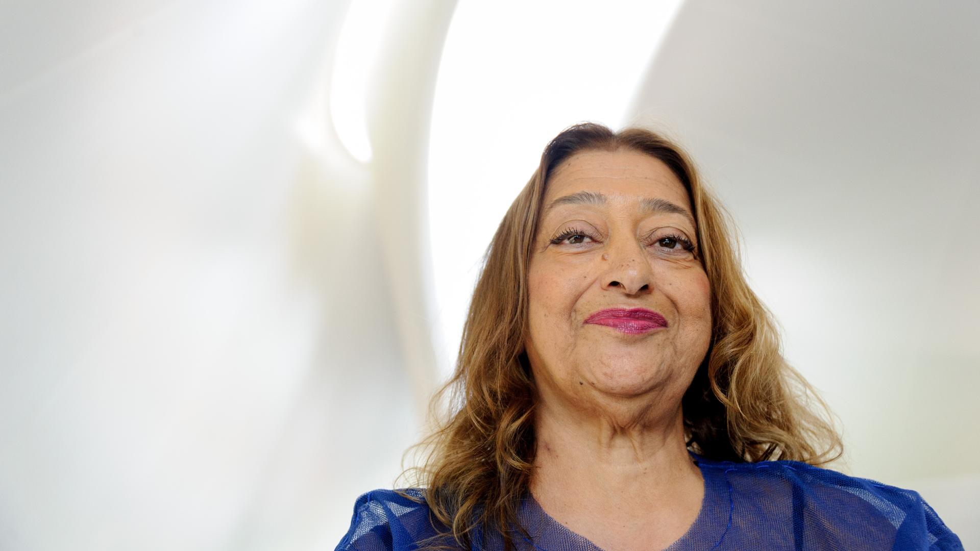 Zaha Hadid lächelt in die Kamera, über ihr fällt Licht durch eine sanft geschwungene Fensteröffnung in den weißen Raum.